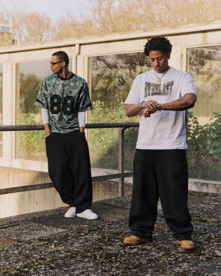 To mænd poserer udendørs i urban omgivelser med oversize streetwear. Den ene bærer en camouflagemønstret Everlast trøje med tallet "88" og hvide sneakers, mens den anden har en hvid Everlast t-shirt med grønt logo og kigger på sit ur iført brune støvler. Baggrunden består af beton, glas og efterårsfarvede træer.