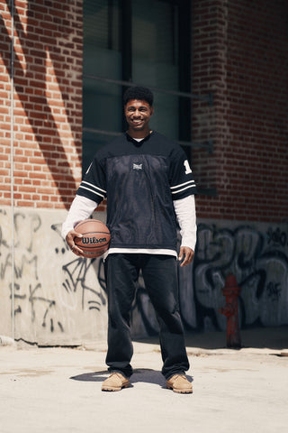 En mand står selvsikkert udendørs med en Wilson-basketbold i hånden, iført en sort Everlast-trøje over en hvid langærmet skjorte, sorte jeans og brune sneakers. Han poserer foran en murstensvæg dækket af graffiti og blander atletisk stil med streets.