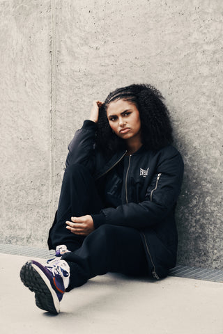 En kvinde med krøllet hår sidder på jorden op ad en betonvæg, iført en sort Everlast bomberjakke, sorte bukser og lilla sneakers. Hendes udtryk er alvorligt, og den urbane omgivelse komplementerer det streetwear-inspirerede outfit.
