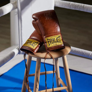 Et par vintage-inspirerede brune Everlast-boksehandsker med gule mærker ligger på en træskammel inde i en boksering. Den blå måtte og de hvide reb indrammer scenen og fremkalder et klassisk trænings- eller kampmiljø.