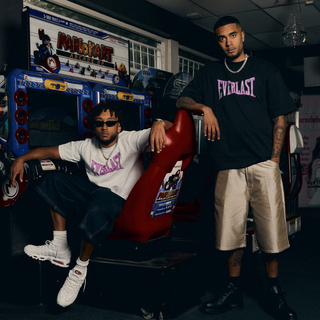 To mænd, Oliver Kesi og Benny Jamz, poserer foran et arkadespil, iført oversized Everlast-T-shirts med fed pink skrift. Den ene sidder i en rød arkadestol iført en hvid skjorte, solbriller og hvide sneakers. mens den anden står ved siden af ham i en sort t-shirt og beige shorts. Retro arkaden baggrunden tilføjer en nostalgisk streetwear vibe.