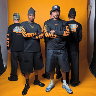 Fem mænd poserer foran en lys orange baggrund iført matchende sort streetwear med markante orange "EVERLAST"-bogstaver på ærmerne og store tal på ryggen. To mænd står med armene over kors og kigger fremad, mens de tre andre vender ryggen til og viser de grafiske designs frem. Scenen blander urban stil med markant branding og en selvsikker gruppedynamik.